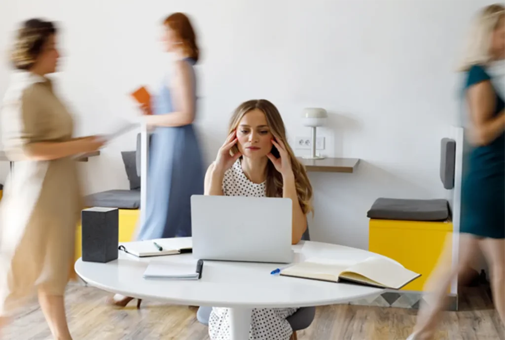 A stressed woman sitting at a laptop with blurred figures walking past, representing the overwhelm and isolation Christian childcare leaders often feel without support.